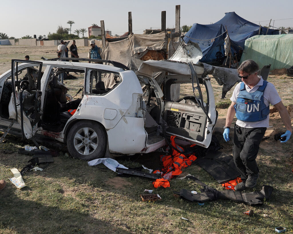 Men in vests that say "UN" and "OCHA" walking near a damaged white car.