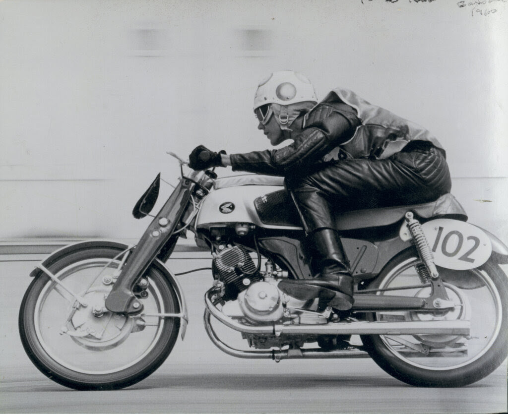 A black and white photo of Mary McGee, in a polka-dot helmet, crouched on a racing motorbike with speed blur behind her.