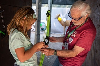 An older, white-haired man in glasses helps a young red-haired girl load an arrow into a bow