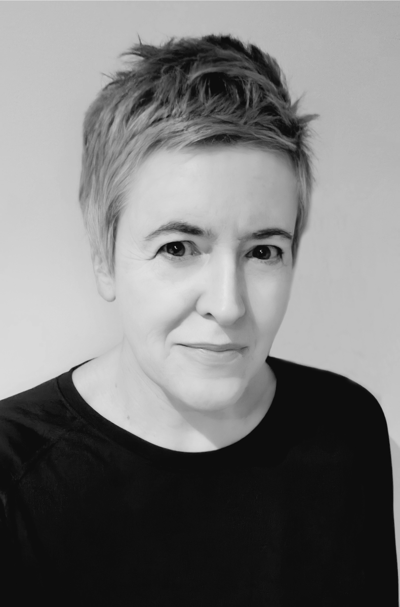 In this black-and-white image, writer Julie Breathnach-Banwait stands in front of a white wall, wearing a black t-shirt.