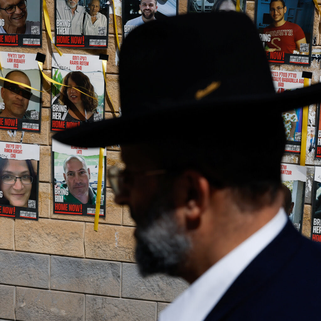 Posters of missing hostages displayed on brown wall. In the foreground is a person in a black hat and glasses.