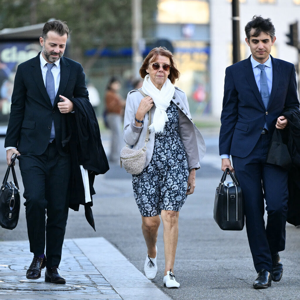 Gisèle Pelicot, wearing sunglasses and a printed dress, walks down the street between two men in suits.