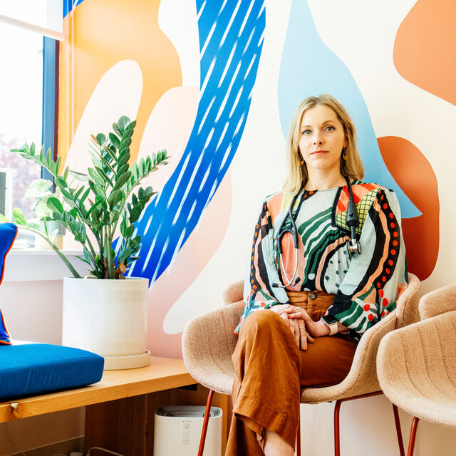 Dr. Stefie Deeds sits in a colorful modern chair in a waiting room of her private practice, which has bright colors and shapes on the walls that are echoed in her brightly colored long-sleeve blouse.