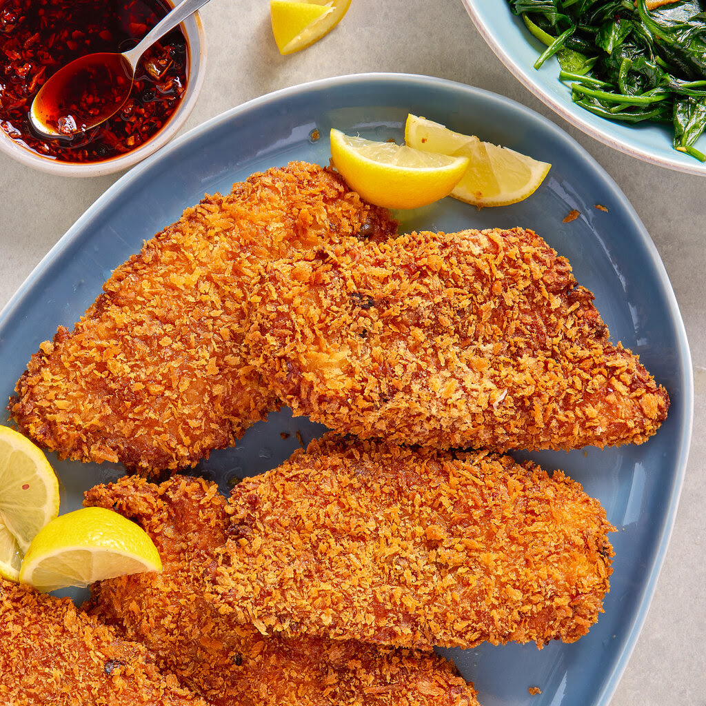 Fried chicken cutlets are on a blue plate with lemon wedges. Next to the plate are dishes with greens and a red, peppery sauce.
