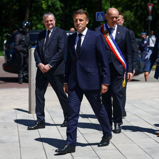 President Emmanuel Macron, in a navy suit, walking outside. His wife, Brigitte, and four men walk behind him.
