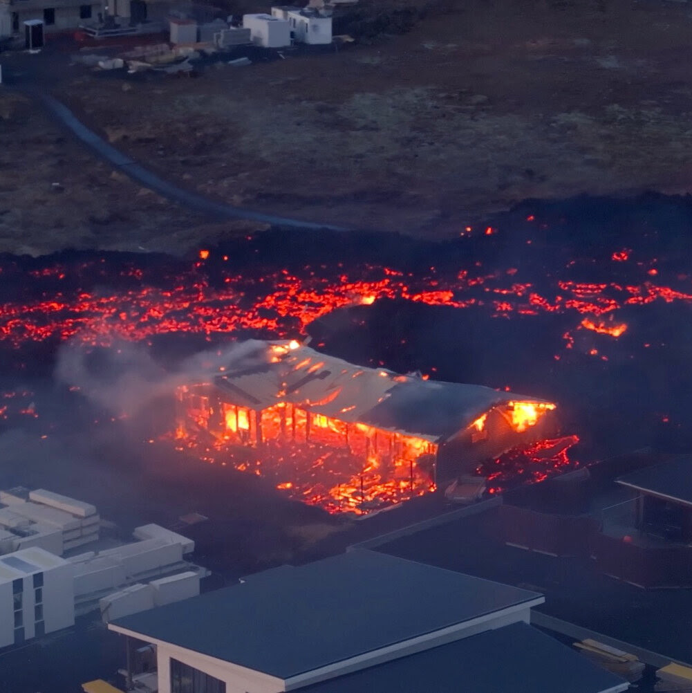 A black smoldering stream of lava overwhelms a house whose interior is in flames. 