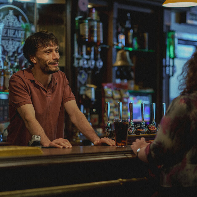 A man stands behind a bar and a woman sits facing him.