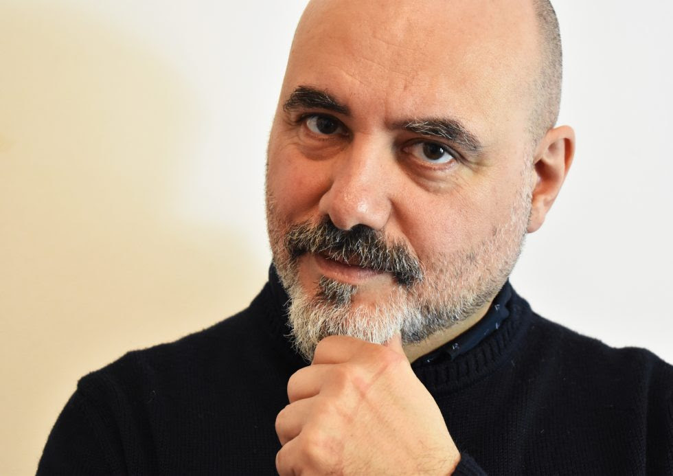 Fabio Banfo, wearing a black sweatshirt, gives the camera an attentive look, his hand raised to his bearded chin. Behind him is a cream wall.