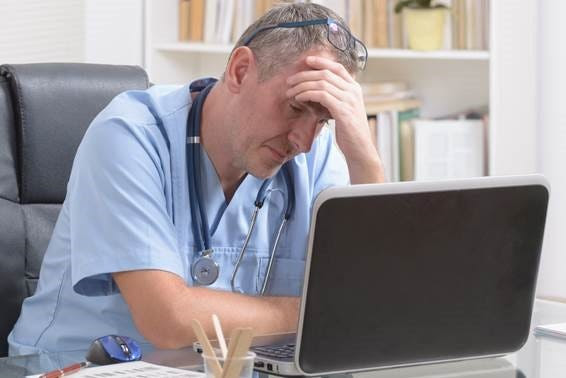 A doctor seated at his desk, focused on his laptop, surrounded by medical books and a stethoscope.