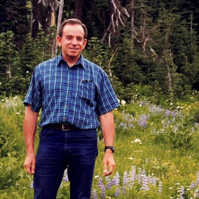 A man in a blue collared shirt and jeans posing for a photo in a grassy field. 