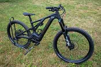 a black e-bike with a gray seat and fat, knobby tires is parked in a back-tire bike rack on a grassy area