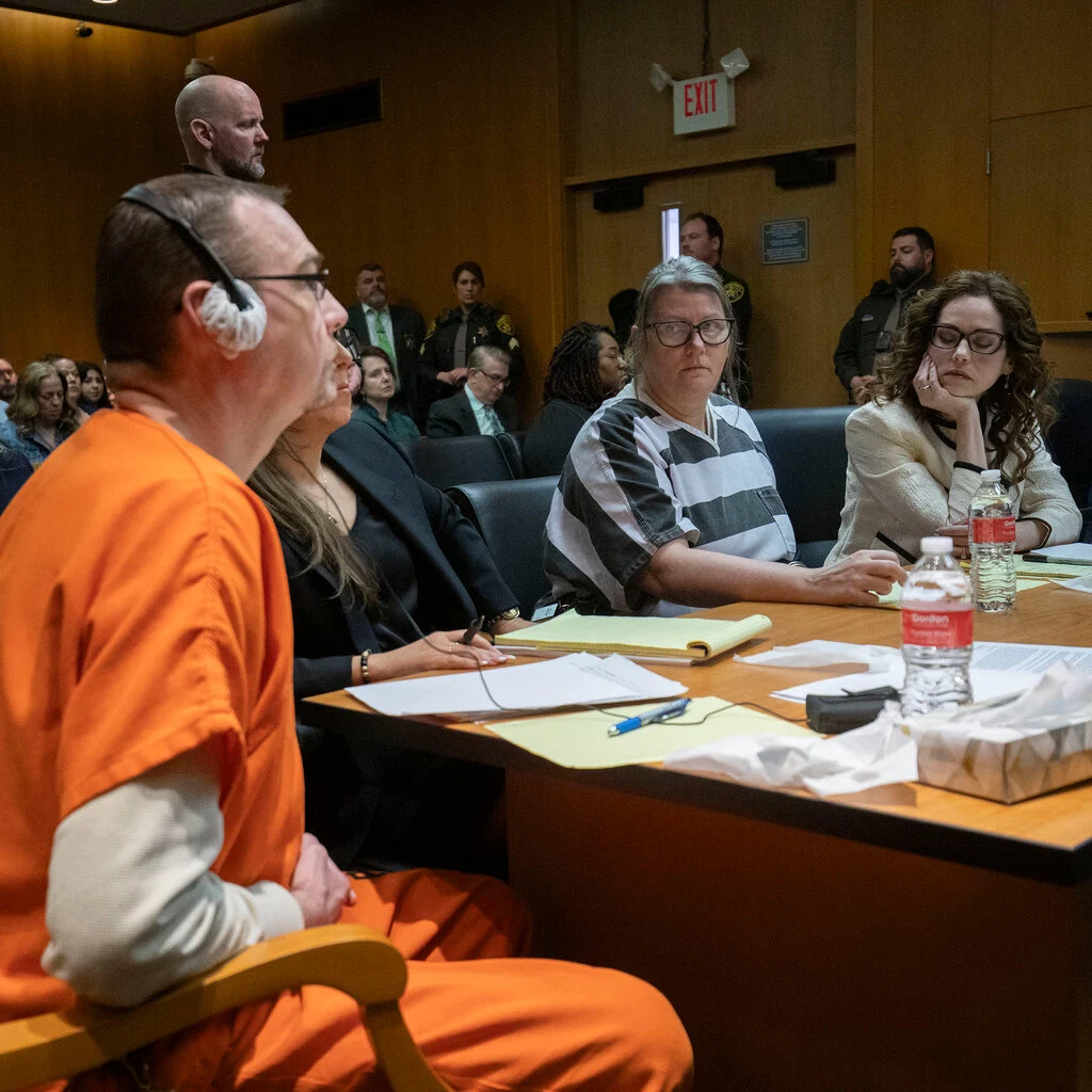 People sit around a table in a courtroom. The man, closest to the camera, wears an orange uniform. A woman across the table, wearing a black and white striped uniform, stares at him. 