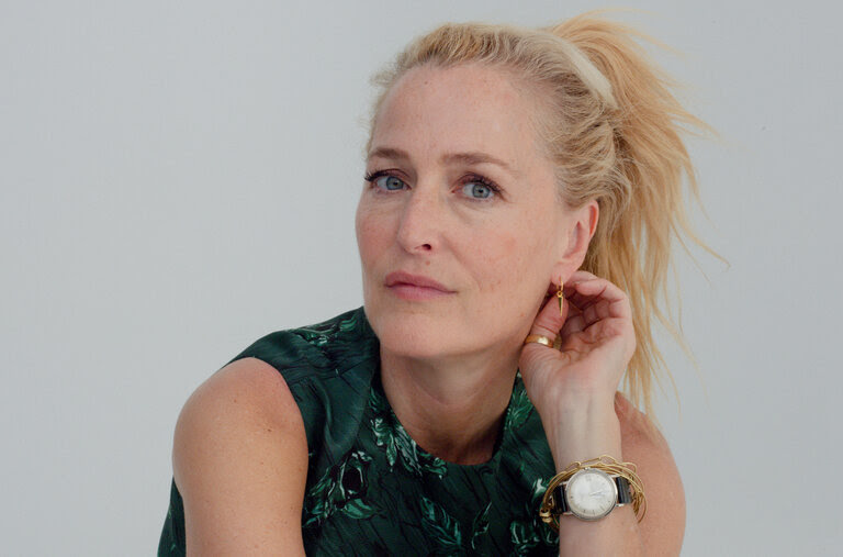 A portrait of Gillian Anderson sitting in a green dress as her hand plays with her earring. She wears a watch and bracelets on her wrist.