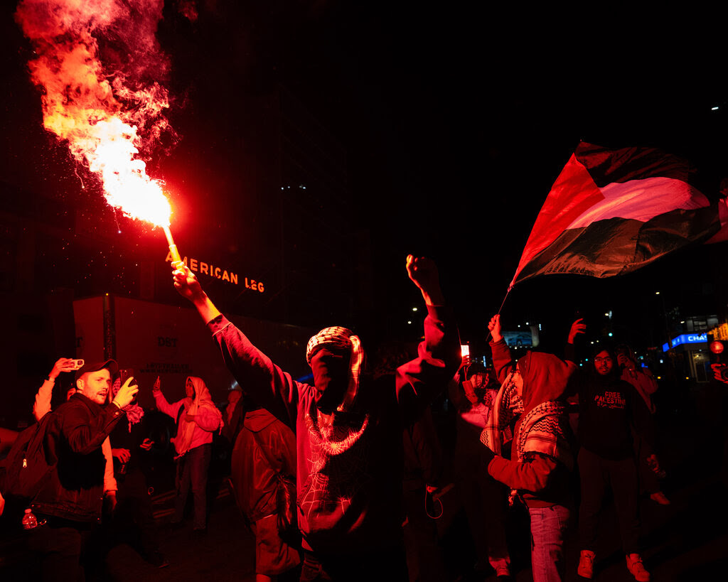 A group of people are bathed in red light from a flare being held up by one person. Another person waves a Palestinian flag.