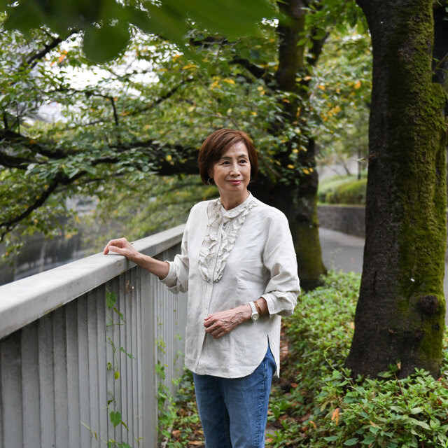 Hideko Yamashita, con una camisa blanca y un reloj y una pulsera en el brazo izquierdo, de pie con la mano derecha apoyada en una valla.