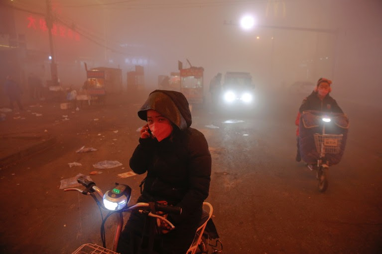 A woman wearing a face mask amongst heavy smog looks to camera while sitting on her bike and speaking on a mobile phone