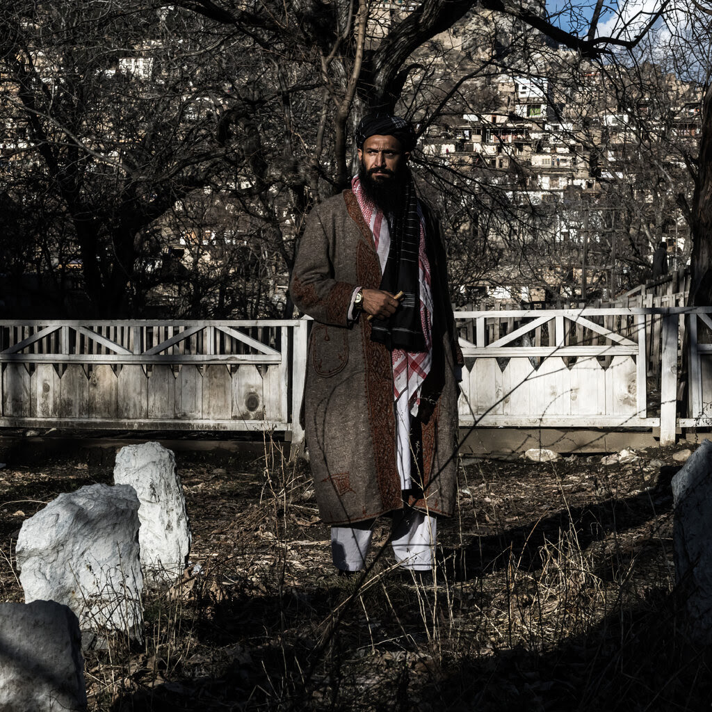 A portrait of Mullah Osman, partly shadowed, standing between white rough gravestones.