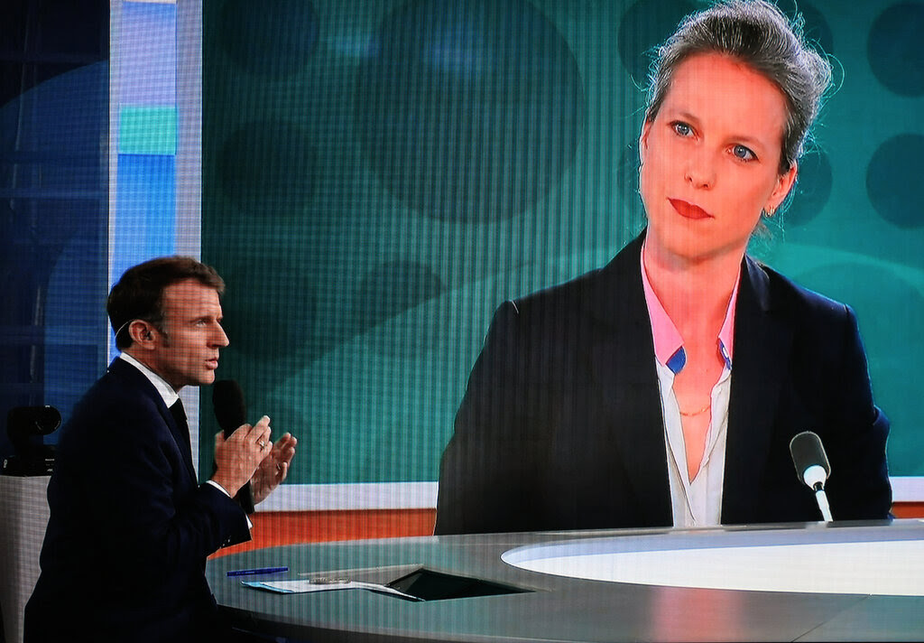 A woman’s image is shown on a giant screen as Emmanuel Macron sits at a round table in a TV studio, gesturing.