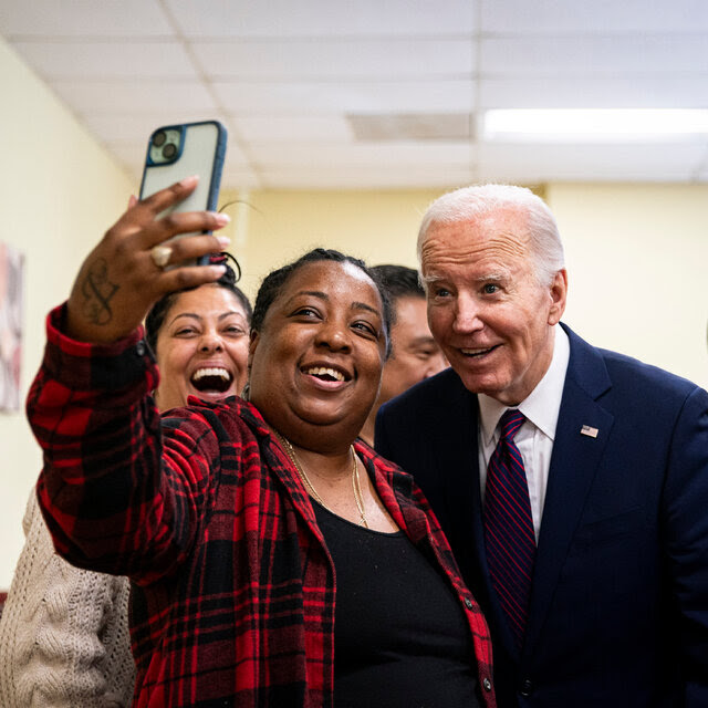 President Biden, in a dark blue suit and tie with red stripes, posing for a selfie with a woman in a black-and-red checked shirt. 