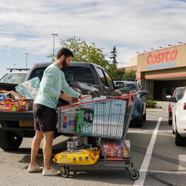 With a Costco warehouse in the background, a man in shorts and a long-sleeved shirt loads groceries from a cart to the bed of a pickup truck.