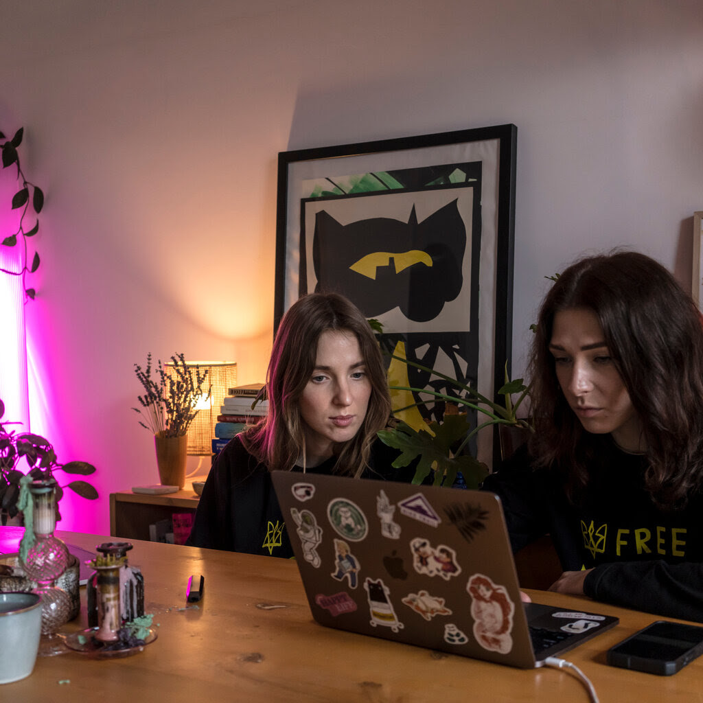 Two women sit at a table looking at a laptop computer, with plants and artwork in the background.