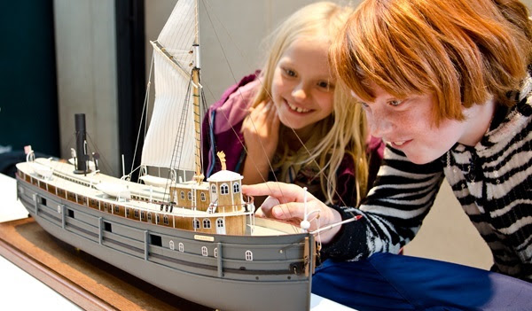 young girl and boy smile, wide-eyed, as they look at a model ship with a masted sail