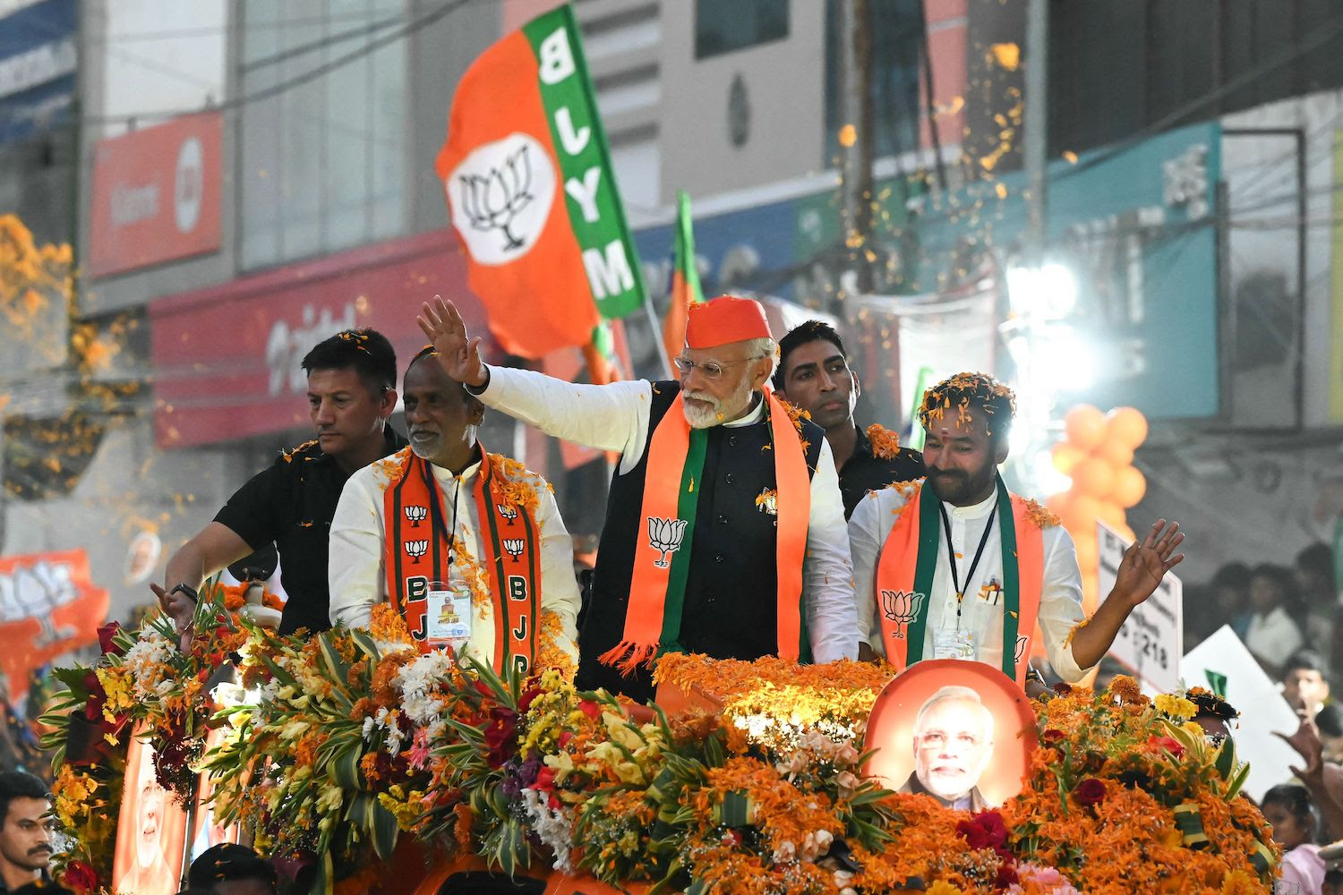 Indian Prime Minister Narendra Modi waves to the crowd as he stands in a float during a campaign event. Modi wears a black vest with a scarf and hat in the orange-and-green colors of the Indian flag. The float is also decorated with orange flowers and photos of Modi, and he is flanked by security officers and other members of his party.