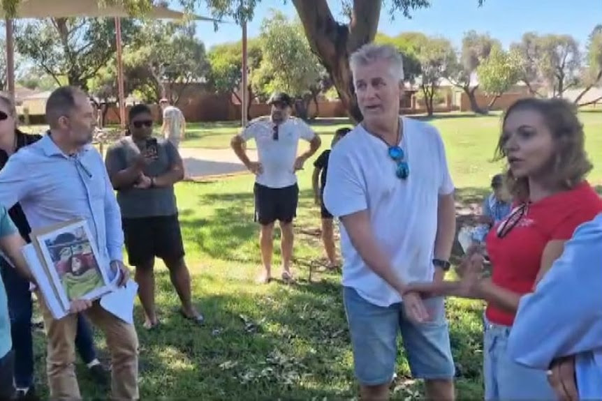 A woman in a red t-shirt speaks to a group of people in a park. One of the men in the group holds a picture.