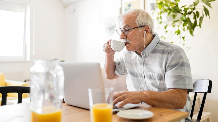 An older adult drinks coffee while at their computer.