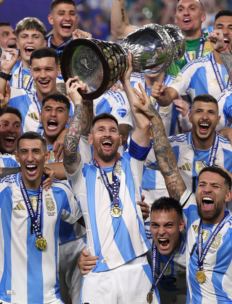 The Argentina soccer team celebrates on the field. Lionel Messi hoists a trophy cup over his head while standing in the middle of the team as they crowd together. 