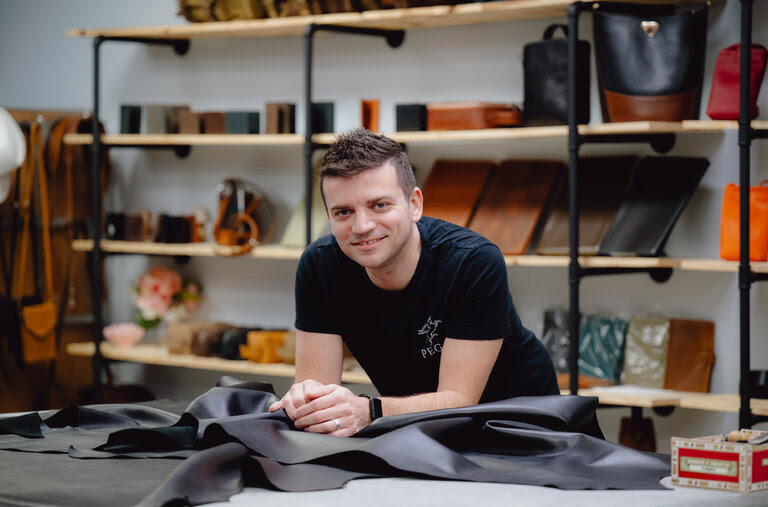 Mr. Yilmaz, in a black T-shirt, is seen in his store, smiling and leaning over a counter spread with black leather material. Behind him is shelving displaying bags, wallets and other leather goods.