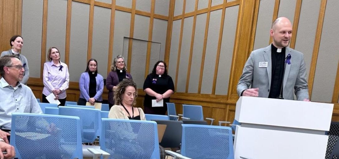 Rev. Tim Schaefer speaks at a white podium during a Madison Common Council meeting. Standing along the wall behind him are Rev. Breanna Illéné, Linda Ketcham, Rev. Gayle Tucker, Rev. Cathy Weigand, and Rev. Mallory Yanchus.