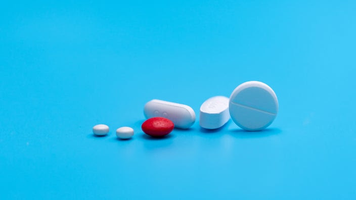 A group of round and oval white tablets pills as well as a red sugar-coated tablet are pictured against a blue background.