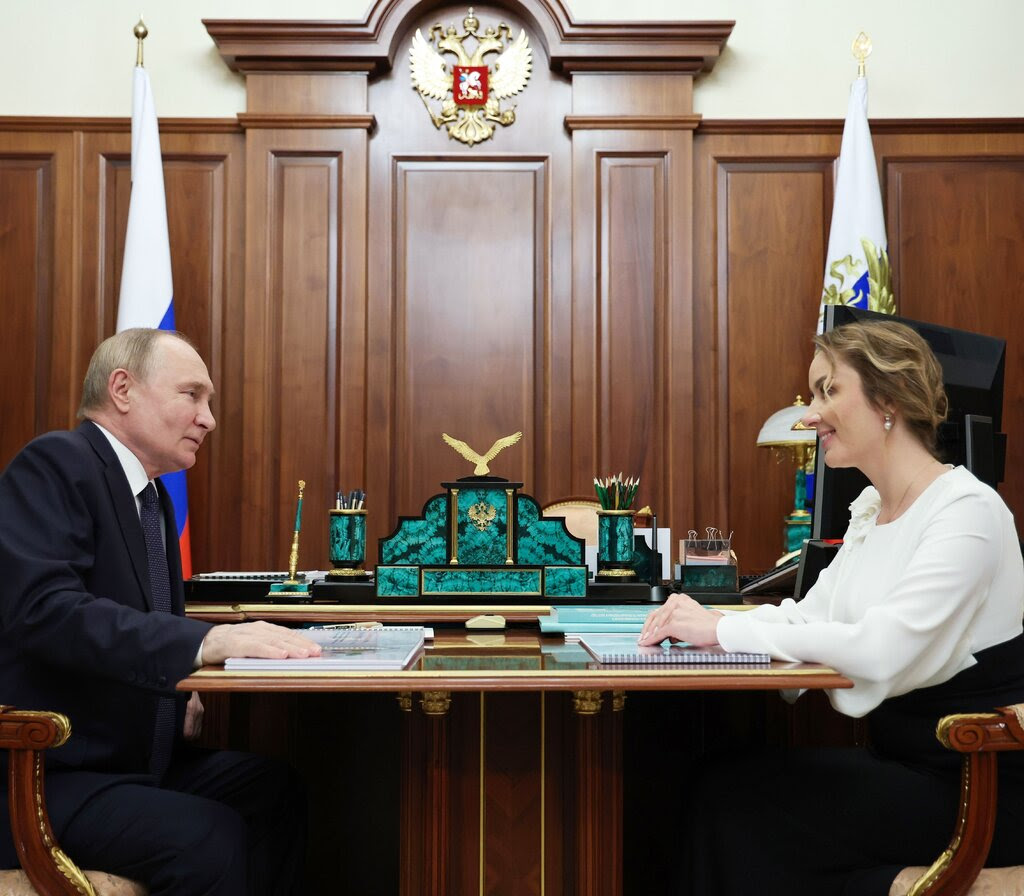 Vladimir Putin and Maria Lvova-Belova sit opposite each other at a table beside a wall with wooden paneling.