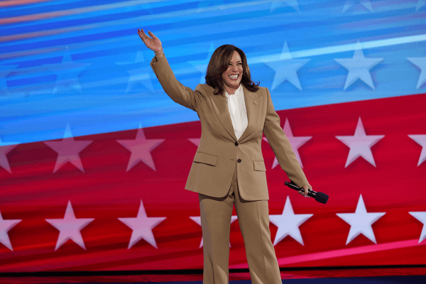 Vice President, Kamala Harris, waves and smiles on a stage holding a microphone and a tan suit. Foreground is red, white, and blue. 