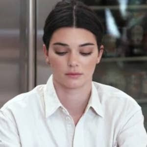 Person with hair tied back wearing a button-up shirt is looking down, seated in a kitchen setting