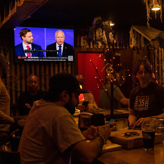 Two televisions showing the debate are on the wall of a restaurant. A man at the bar is looking at his phone.