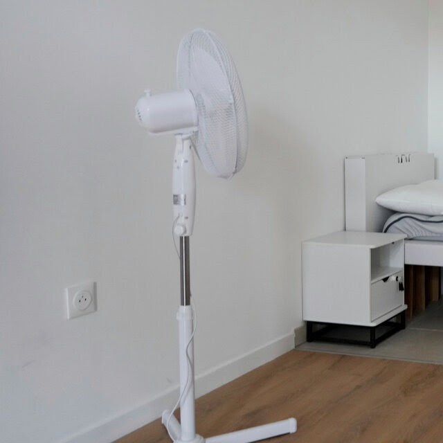 A small bedroom has white walls and a wooden floor. A white bed is in the corner and there is a white fan on a stand near the wall. 