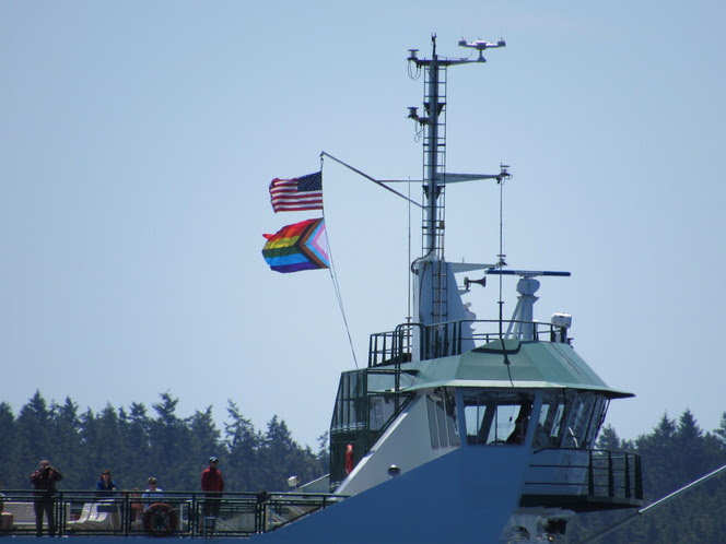 Ferry bridge with American and Pride flags, radar tower, people on deck and trees in the background.