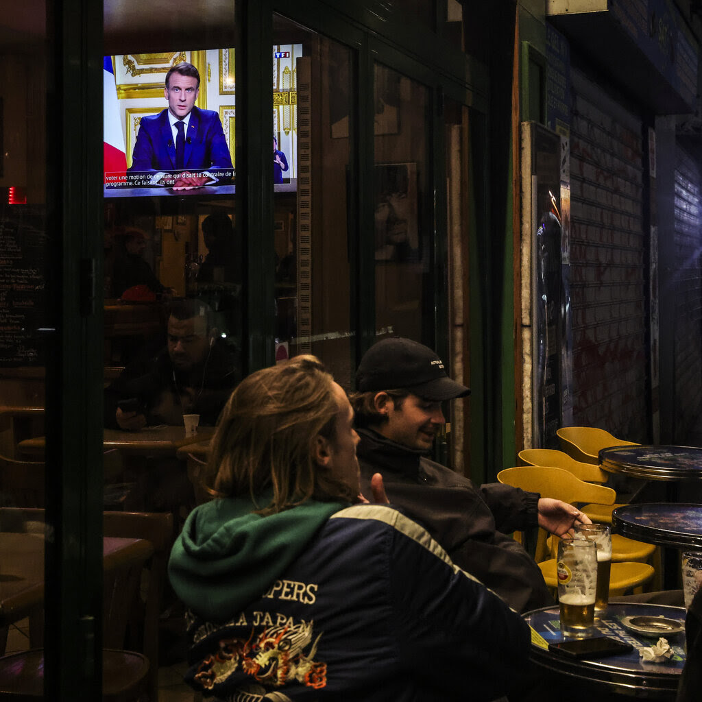 People sit at night outside a bar in which a television screen shows the French president giving a speech.