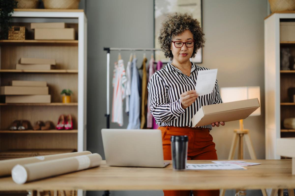 Young online merchant woman writing information and address on parcel box to preparing clothes delivery for customer while working