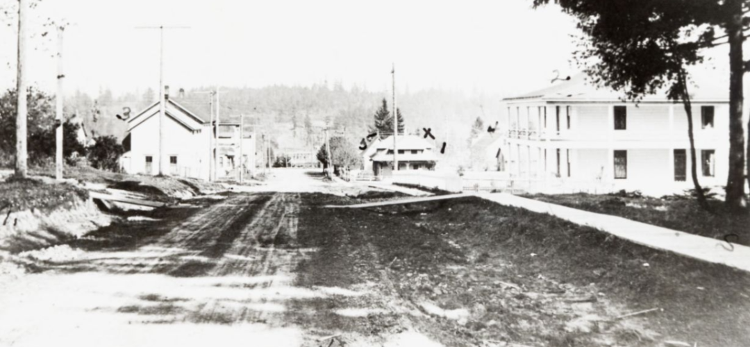 A historic photo from 1908, black and white. there are a few white buildings visible, a hill in the distance, and a dirt road