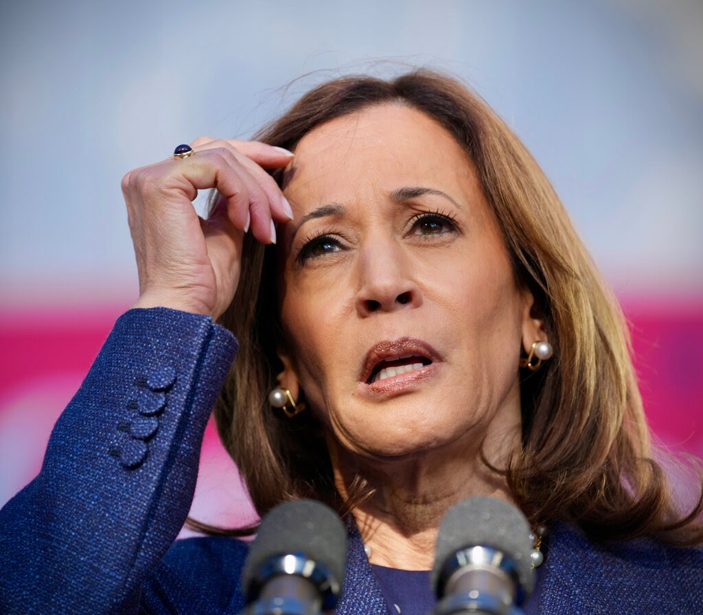 Kamala Harris holds her right hand to her forehead while speaking into microphones.