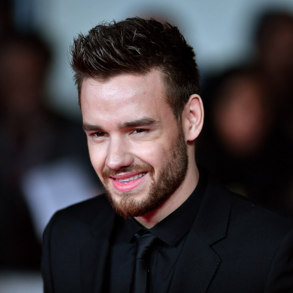 Liam Payne, wearing a black suit, shirt and tie, smiles while looking past the camera.