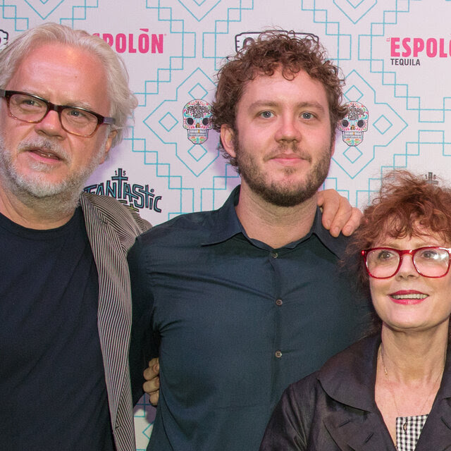 A man in a dark button-up shirt looks toward the camera. On his sides are Tim Robbins, who is wearing a corduroy jacket, dark shirt and glasses, and Susan Sarandon, who is wearing a black leather jacket and red glasses.