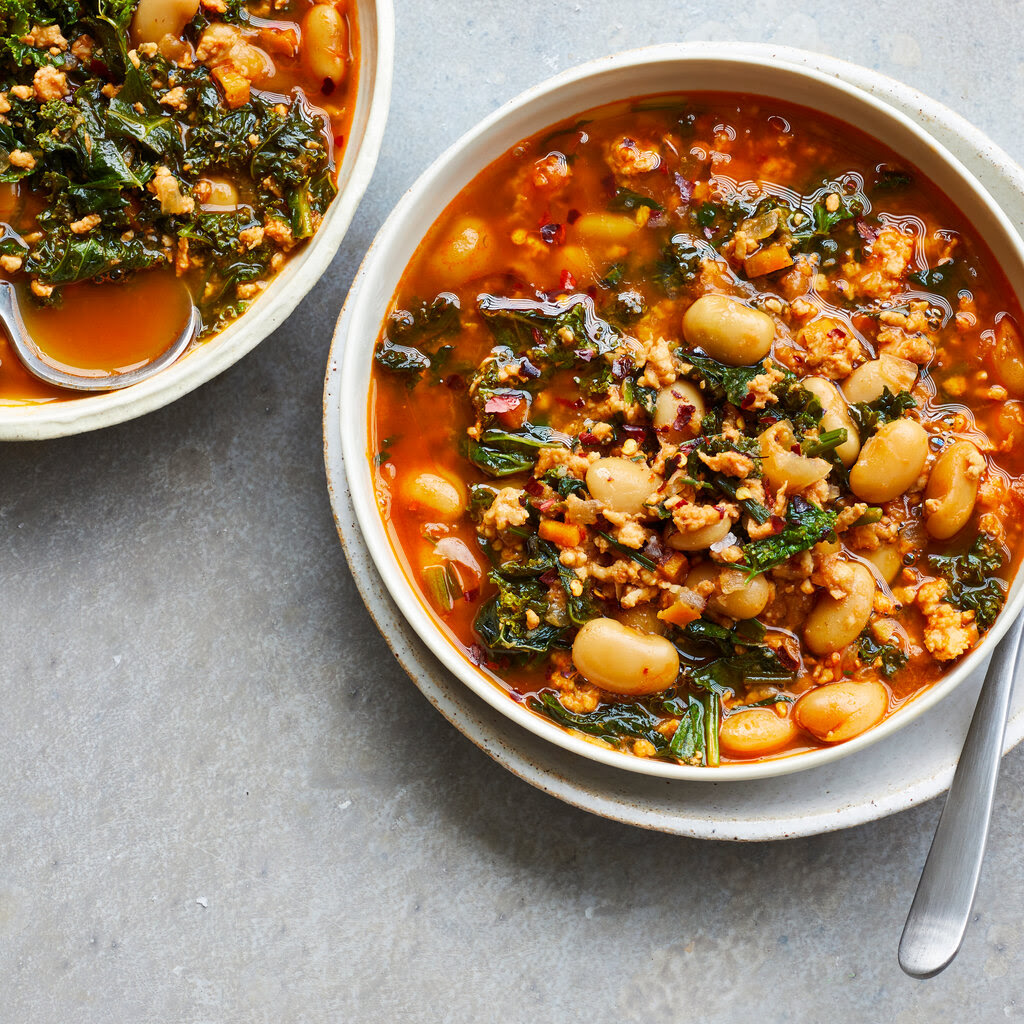 Two white bowls of soup on a gray surface, each with metal spoons. The soup is a red broth filled with greens, white beans and bits of ground turkey.