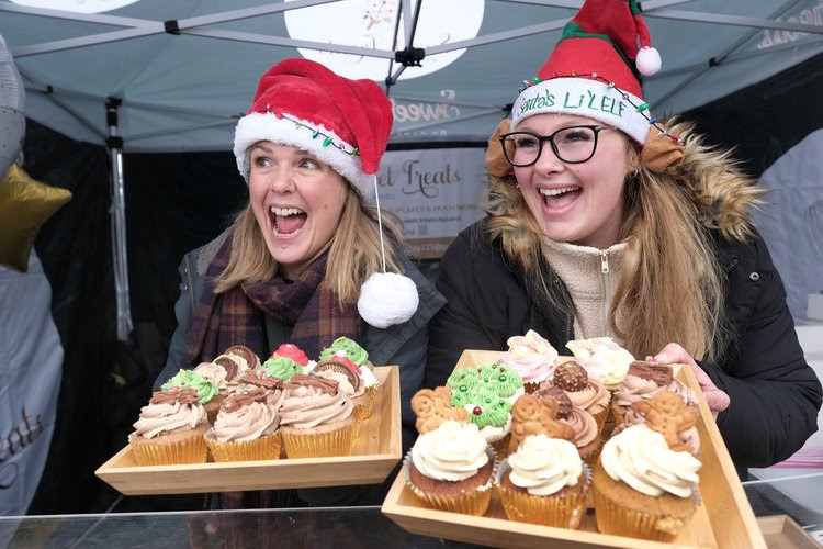 Two smiling stall holders at the Malton Christmas Festival wearing Christmas hats and holding large, festive cupcakes.