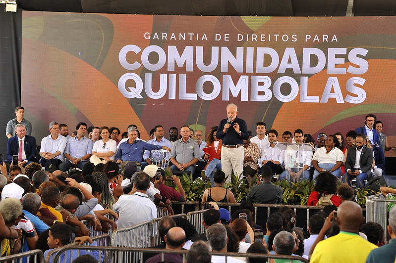 Presidente Lula fala ao centro de palco, com várias autoridades ao lado e plateia cheia