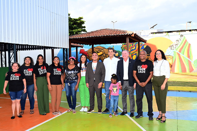 Foto colorida mostra várias pessoas em quadra de escola. Todas estão em pé e sorrindo, na escola da Liberdade, durante entrega de títulos de regularização fundiária.