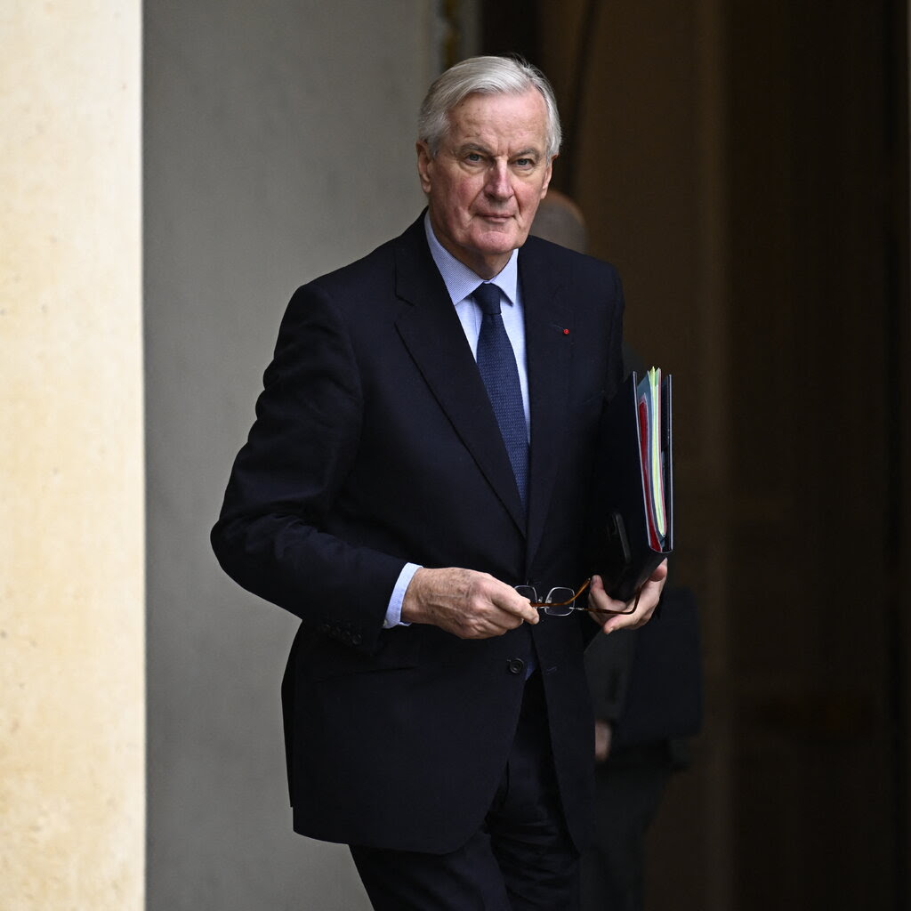 A man holds eyeglasses in his hands. He is wearing a dark suit and a blue tie.